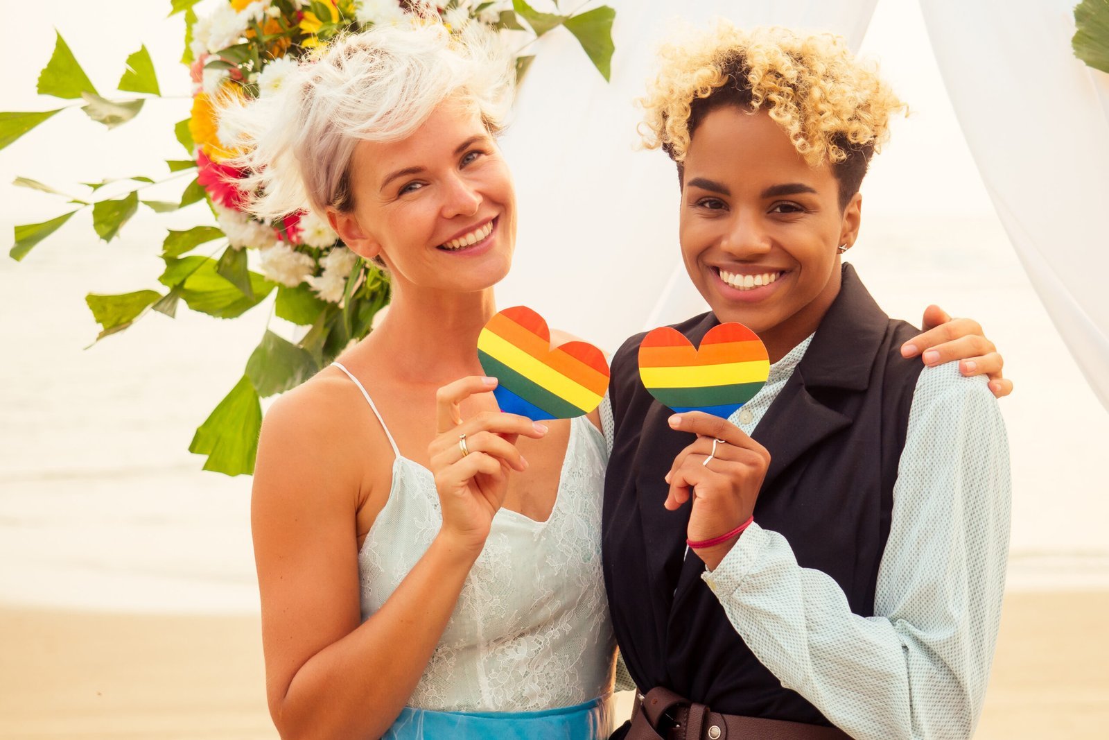 african american female groom in black suit and caucasian bride in dress in ceremony on tropical beach under wedding flover arch